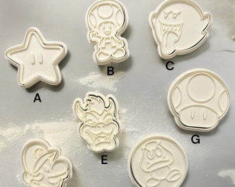 Puede incluir: Conjunto de 7 cortadores de galletas de plástico blanco con forma de personajes de la franquicia de videojuegos Super Mario Bros. Los cortadores están etiquetados de la A a la G.