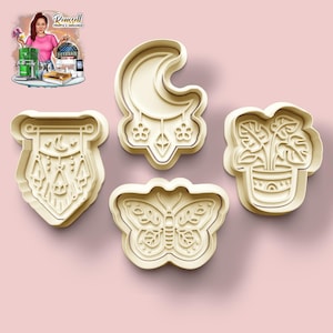Pode incluir: Quatro cortadores de biscoitos bege em forma de lua crescente com pingentes, uma borboleta, uma planta em vaso e um design geométrico com um motivo de apanhador de sonhos. Em um fundo rosa.