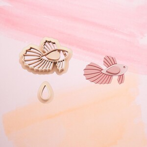 May include: Two wooden cookie cutters in the shape of a betta fish. One cutter is a solid pink color and the other is a detailed outline. A small teardrop-shaped cutter is also included.