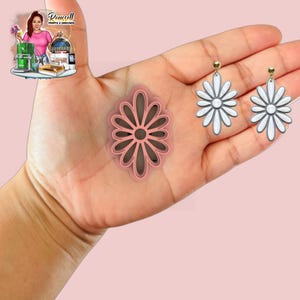 May include: A pink daisy-shaped cookie cutter and two daisy earrings with white petals and grey centres are displayed. The earrings have gold-coloured posts. The items are held in a hand against a pink background. The cookie cutter is oval-shaped.