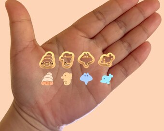 May include: A hand displaying a set of eight gold-colored cookie cutters shaped like various sea creatures, including a crab, a seahorse, a stingray, a dolphin, and a seashell.