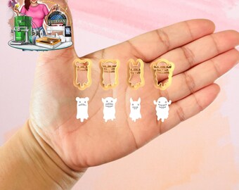 May include: A hand displays several small, yellow cookie cutters in various monster shapes, alongside matching white cookie cutouts. The cutters have detailed designs, and the cutouts are simple, white versions of the same monsters. The background is a soft pink.