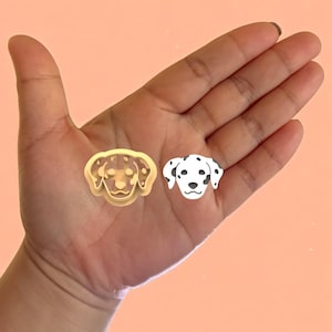 Op de afbeelding: Twee hondenitems. Eén is een goudkleurige koekjesvorm en de andere is een wit-zwarte Dalmatische hondensticker. De items worden op de hand van een persoon weergegeven tegen een perzikkleurige achtergrond.