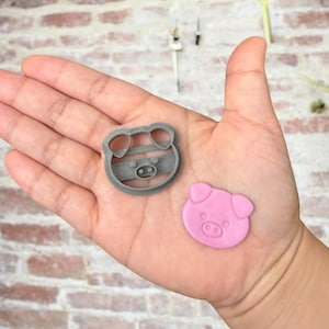 May include: Two pig-shaped cookie cutters, one gray and one pink. The gray cutter has a striped pattern and the pink cutter is solid.