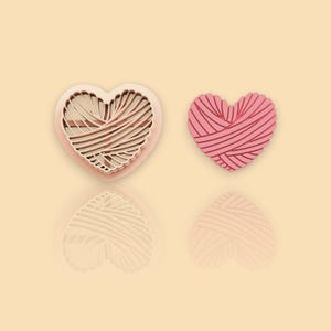 May include: Two heart-shaped cookie cutters, one pink and one beige, with a yarn pattern design.
