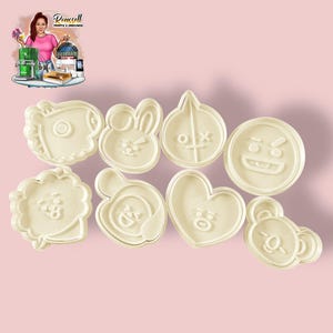 May include: A collection of eight off-white cookie cutters in various cartoon character shapes. The cutters include a bunny, a heart, a bear, and other stylized faces. The cookie cutters are arranged on a light pink background.
