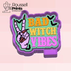 May include: A purple, green, and orange cookie cutter with the text "BAD WITCH VIBES" and a hand making a peace sign with long, red fingernails. The design includes bats. The logo "Roussell Prints" is in the upper left corner.