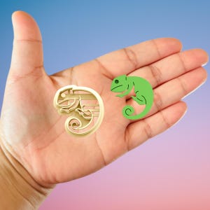 May include: A gold-colored chameleon-shaped cookie cutter next to a green chameleon sticker. The cookie cutter has an ornate design, while the sticker is a simple, cartoon-like illustration. Both are displayed against a gradient background.