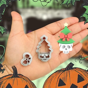 May include: Three gray plastic cookie cutters shaped like Christmas trees with skulls on the bottom. The trees have a star on top and a red bow. The cutters are on a hand with a pumpkin in the background.
