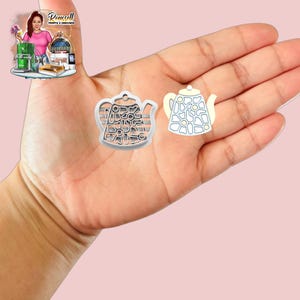 Può includere: Due ciondoli a forma di teiera. Uno è color argento con un design traforato, l'altro è bianco con un motivo floreale blu. I ciondoli sono tenuti in una mano su uno sfondo rosa. L'immagine include anche una piccola illustrazione di una donna.