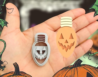 May include: Two light bulb shaped cookie cutters, one grey and one orange, with a jack-o'-lantern face design. The cutters are held in a hand against a background of green vines and pumpkins.