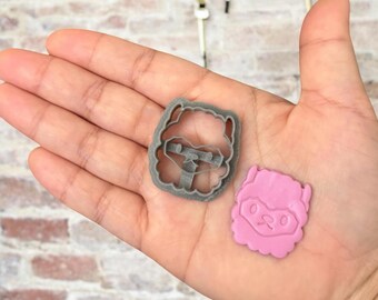 May include: A gray plastic llama cookie cutter and a pink llama cookie. The cookie cutter is in the shape of a llama's head with a fluffy mane and ears. The cookie is a similar shape and has a pink glaze.