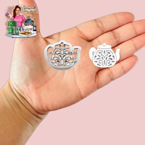 Può includere: Due formine bianche a forma di teiera. La formina più grande presenta un elaborato motivo floreale all'interno del suo contorno. La formina più piccola è una versione in miniatura con un motivo floreale simile. Entrambe sono tenute in mano su sfondo rosa.