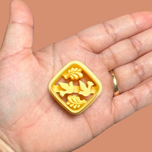 Puede incluir: Un cortador de galletas de plástico amarillo en forma de diamante con dos palomas y hojas en su interior.