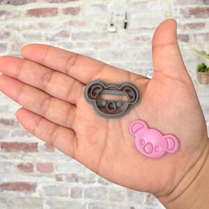 May include: Two koala cookie cutters, one gray and one pink. The gray cutter is a detailed outline of a koala's face, while the pink cutter is a solid shape of a koala's face.