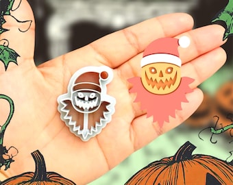 May include: Two Halloween-themed cookie cutters. One is a white plastic cutter in the shape of a scarecrow wearing a Santa hat. The other is a pink and yellow scarecrow with a Santa hat. The background includes a hand and orange pumpkins.