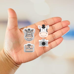 May include: Four small, light-coloured cookie cutters in the shape of a zebra head, a camera, and a zebra face with a camera. The zebra head has a black and white striped pattern. The camera cutters are grey and have a black lens.