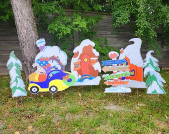 May include: A collection of colorful, whimsical Christmas yard decorations. The set includes a car with a character, a house, directional signs, and snow-covered trees. The decorations are made of a durable material and are designed to be placed in a yard.