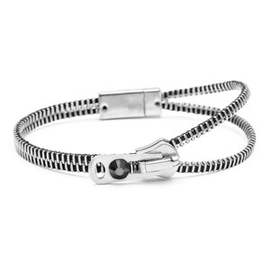May include: A silver-toned bracelet designed to resemble a zip, featuring a metallic slider element with a single dark circular accent. The band consists of two parallel rows of interlocking teeth that meet at a rectangular clasp closure.