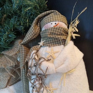 May include: A white snowman with a green and white plaid hat and scarf. The snowman is holding a bundle of dried grass and has seashells and starfish attached to it.