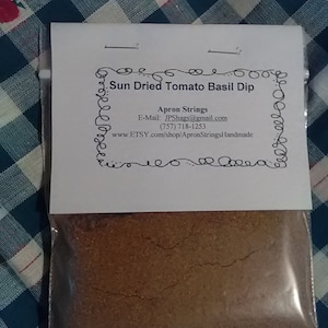 May include: A clear plastic bag with a white label that reads "Sun Dried Tomato Basil Dip" and includes contact information for Apron Strings. The bag contains a brown powder.