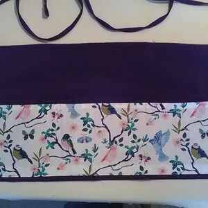 May include: A purple apron with a white pocket featuring a floral pattern with birds and butterflies. The apron has a long tie that can be tied around the waist.