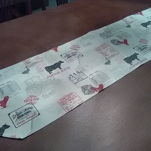 May include: A white table runner with a farmhouse theme. The runner features a black and white print of cows, chickens, and farm equipment. The text "Fresh Eggs", "Green Acres Farm Store", "Premium Quality", and "Farm Fresh Produce Dairy Products" is printed on the runner.