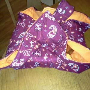 Peut inclure: Un sac fourre-tout en tissu marron et orange avec un motif cachemire et les lettres "VT" répétées sur toute la surface. Le sac est plié et posé sur une surface en bois.