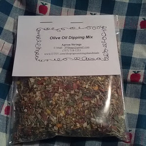 May include: A small white bag with a label that reads "Olive Oil Dipping Mix" and includes contact information for Apron Strings. The bag contains a blend of dried herbs and spices.