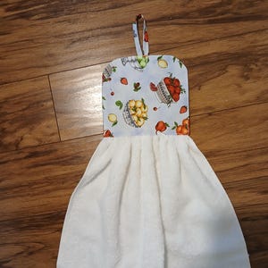 May include: A white kitchen towel with a loop for hanging. The top portion features a light blue background with illustrations of fruit baskets, lemons, strawberries, and peaches. The towel is made of a soft, absorbent material.