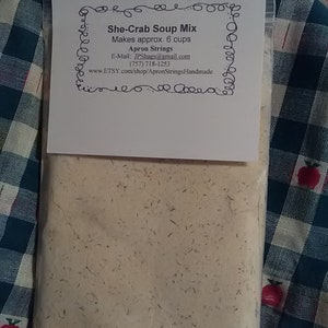 May include: A clear plastic bag filled with a beige powder labeled "She-Crab Soup Mix" with instructions to make approximately 6 cups. The bag is on a blue and white checkered fabric.