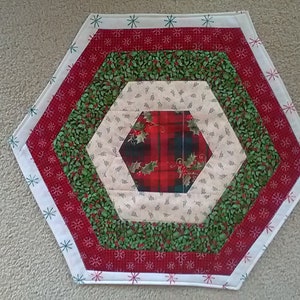 May include: A hexagonal table runner with a Christmas theme. The center features a red and green plaid fabric surrounded by layers of green holly, cream with a small floral pattern, and red with a snowflake pattern. The outer border is white with a blue snowflake pattern.