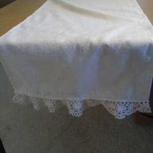 May include: A white table runner with a lace trim. The runner is made of a soft, woven fabric and has a subtle texture.
