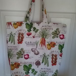 May include: A white canvas tote bag with a colorful print of various vegetables, including tomatoes, carrots, radishes, garlic, and artichokes. The text on the bag reads "Heritage Carrots", "Garlic Delicious", "Beet", "Radishes", and "Tomato".