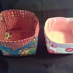 May include: Two fabric storage baskets. One is patterned with red and white checkered fabric, blue fabric with food graphics, and red floral fabric. The other is white with pink lips and a pink interior.