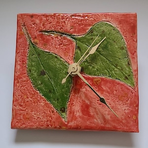 May include: Square ceramic wall clock with a red background and two green leaves. The clock hands and the clock's center are gold-colored. The clock is a decorative item.