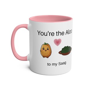Könnte beinhalten: Eine weiße Keramiktasse mit rosafarbenem Rand. Die Tasse zeigt eine Karikatur einer Kartoffel mit einem Herzen und eine Karikatur einer Schüssel mit grünem Blattgemüse. Der Text auf der Tasse lautet "You're the Aloo to my Saag".
