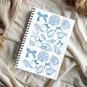 May include: Spiral-bound notebook featuring a blue and white striped pattern. The cover is adorned with blue illustrations of seashells, starfish, and bows. The notebook is open, revealing blank pages.