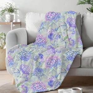 Peut inclure: Une couverture décorative avec un motif floral d'hortensias bleus et violets sur un fond vert clair. Les fleurs sont accompagnées de feuilles vertes. La couverture est posée sur un canapé gris et semble douce et confortable.
