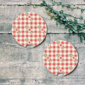 May include: Two round car coasters with a red and cream gingham pattern and small floral accents. The coasters have a cutout for easy removal and are designed to fit in a car's cup holder.