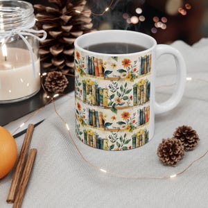 May include: White ceramic mug featuring a colourful design of books on shelves with flowers. The mug is filled with coffee.