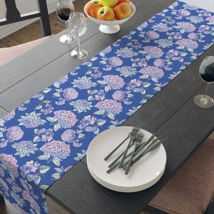 Könnte beinhalten: Ein blauer Tischläufer mit einem floralen Muster aus rosa und weißen Hortensien. Der Läufer liegt auf einem dunklen Holztisch mit einem weißen Teller und Besteck.