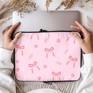 May include: A pink laptop sleeve with a black zipper and a repeating pattern of pink bows and white flowers.
