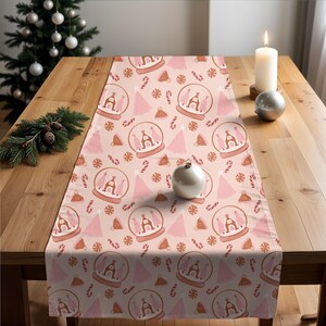 Puede incluir: Un camino de mesa navideño rosa con un patrón repetido de bolas de nieve, árboles de Navidad, copos de nieve y bastones de caramelo. Las bolas de nieve presentan la letra "A". El camino de mesa está sobre una mesa de madera.