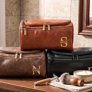 May include: Three personalised leather wash bags in brown, dark brown, and black. Each bag has a gold zip and a gold monogram with a name. A shaving kit with a brush, razor, and bowl of cream sits in front of the black bag.