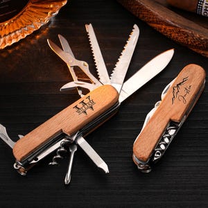 Könnte beinhalten: Zwei Schweizer Taschenmesser aus Holz mit verschiedenen Werkzeugen, darunter Scheren, Sägen und Klingen. Ein Messer ist geöffnet und zeigt seine Werkzeuge, das andere ist geschlossen. Die Messer sind mit den Namen "William" und "Justin" graviert.