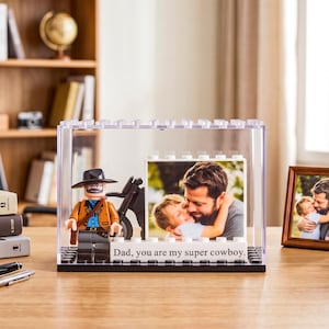 Puede incluir: Un marco de fotos de acrílico transparente con una base negra, con una figura de Lego vaquero y una foto de un padre y un niño. El marco tiene el texto "Papá, eres mi supervaquero". Una foto enmarcada y libros están en el fondo.