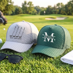 May include: A light grey golf cap with a black graphic of four golf clubs and the text BEST DAD EVER, and a dark green golf cap with a white monogram featuring the letter M and the name MICHALE, both with a textured dimpled surface.