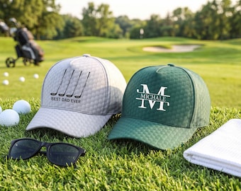 May include: A light gray golf cap with a black graphic of four golf clubs and the text BEST DAD EVER, and a dark green golf cap with a white monogram featuring the letter M and the name MICHALE, both with a textured dimpled surface.