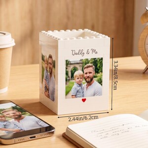 以下が含まれることがあります： 「Daddy & Me」の文字と父と子の写真が入った、白いブロック型のフォトホルダー。ホルダーの高さは8.5cm、幅は6.2cmです。写真の下には赤いハートがあります。